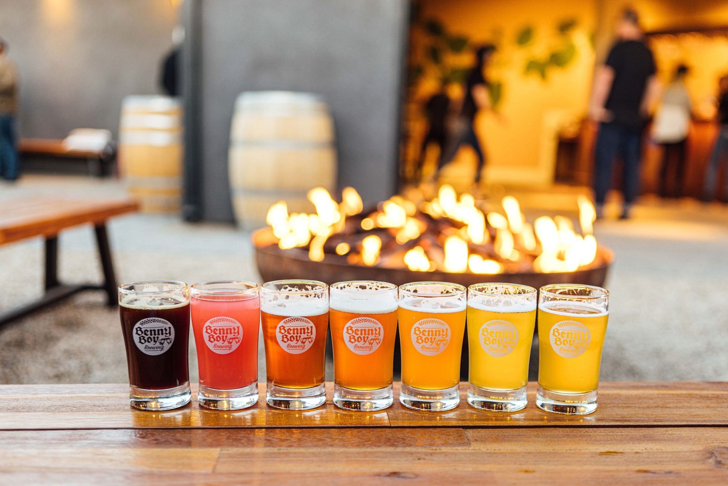 7 color-coordinated beers, darkest to lightest left to right. fireplace in background, wooden barrels