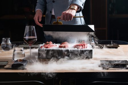 sliced red meat on a black box, a lot of smoke, man in gray suit holding black box top, wine glasses