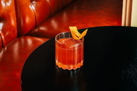 Lowball glass with orange-colored liquid and orange peel twist on a table in front of a red booth seat