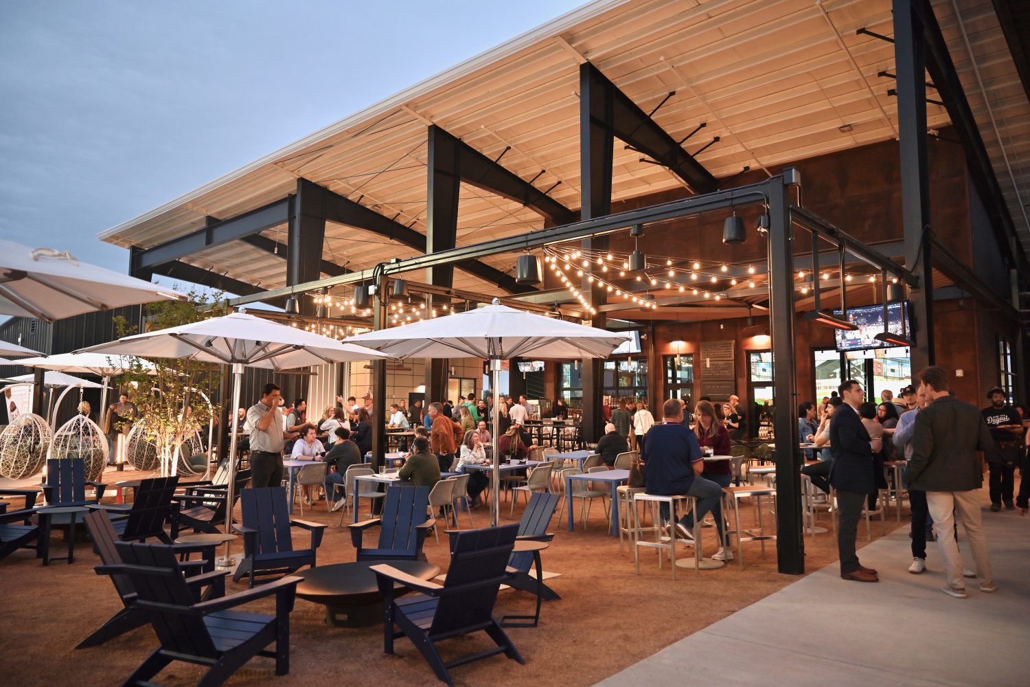 people standing and sitting outdoors drinking, chairs, tables, umbrella, lights, plants