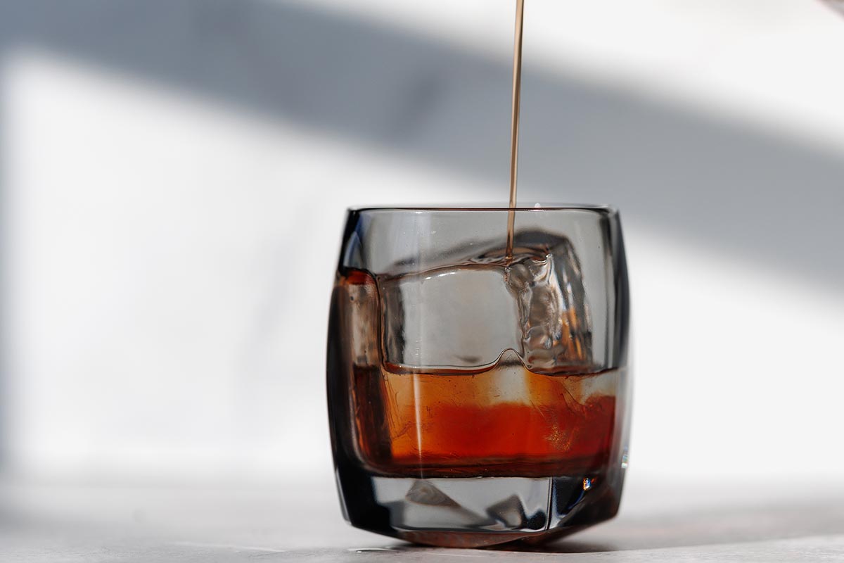 A rocks glass with large ice cube with a drink (off-camera) being poured in