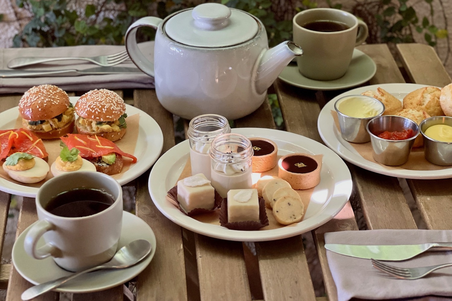 a wooden table with white plates of chicken salad sandwiches, deviled eggs, petit four