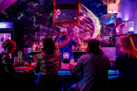 Bartender shaking a cocktail in front of people sitting at a bar in dim lighting with neon- colored disco ball lights