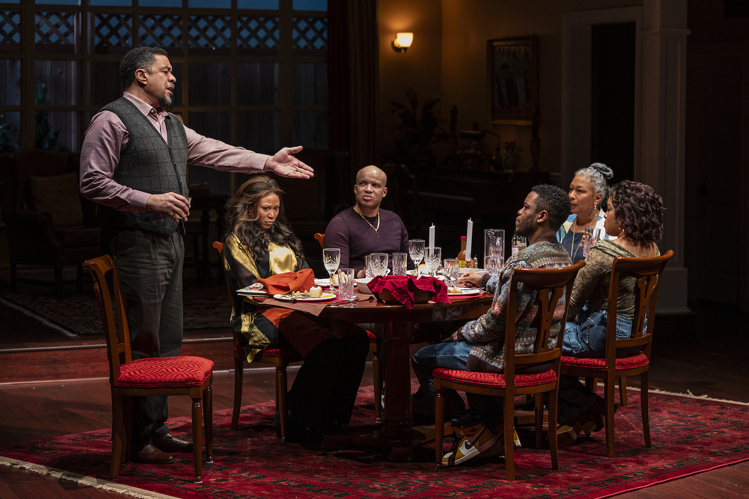 a family sitting around table on stage with one man standing and talking with his hand outstretched