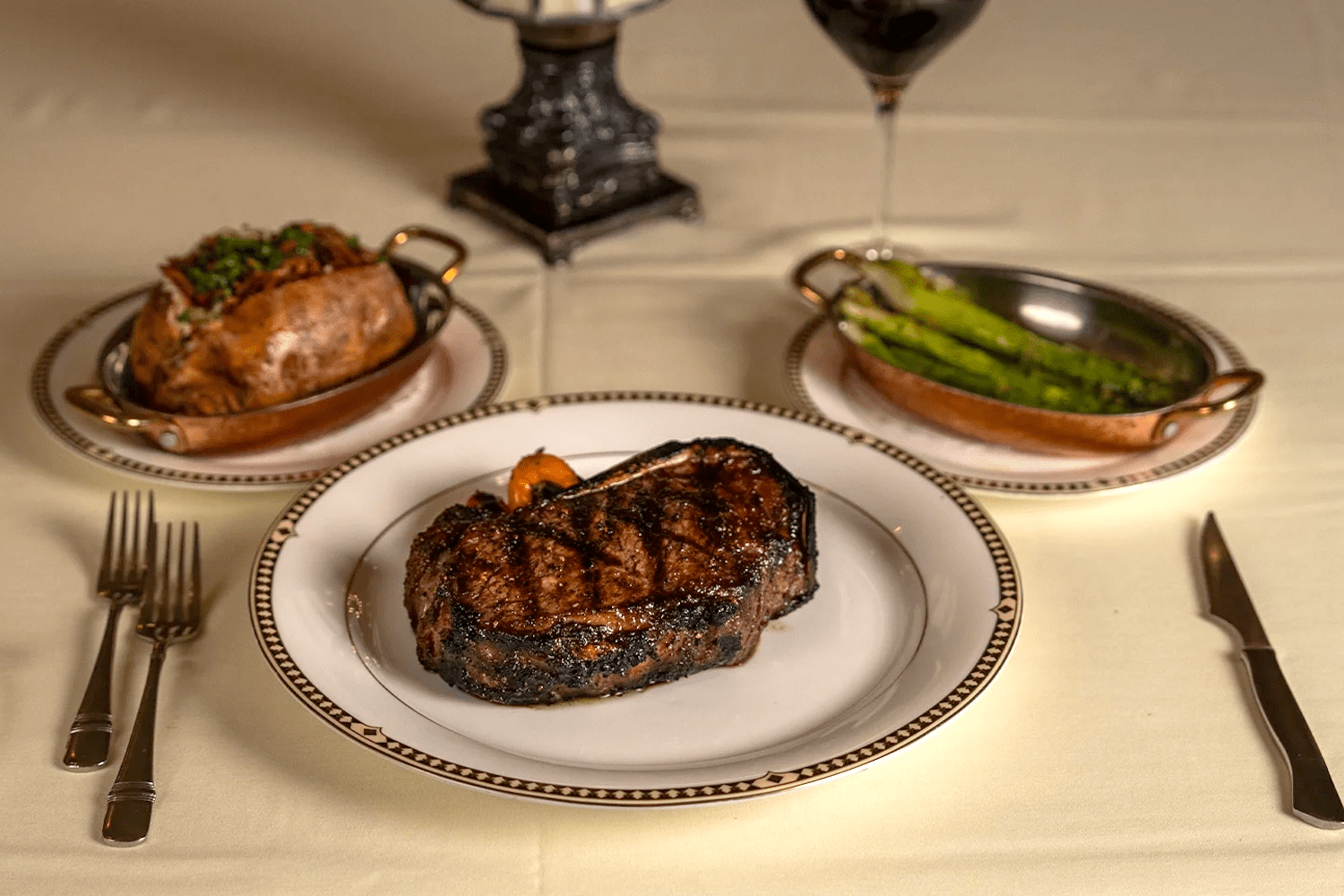 steak on a plate, asparagus on a plate, baked potato on a plate, two forks and one knife