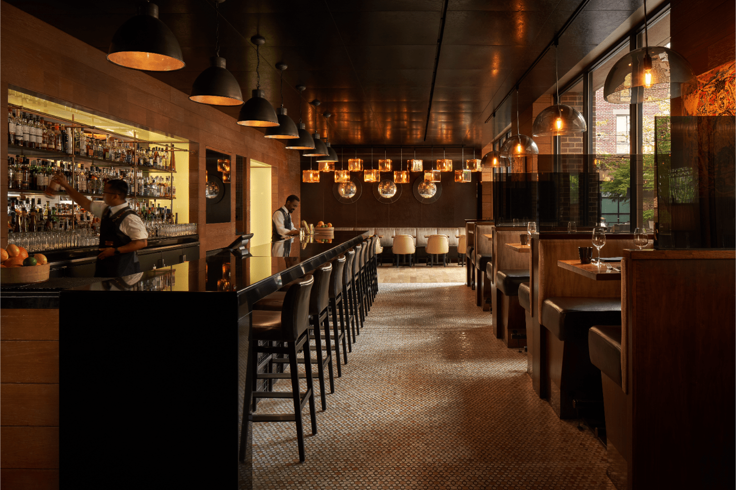 restaurants/bar with brown interior, lamps, liquor on shelves, two bartenders, fruits in a basket, booths, wine glasses on table