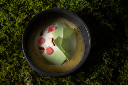 some kind of dessert: yellow bubbly substance, white cream substance, green broken shards, pink and yellow petals, inside black bowl, on top of faux grass