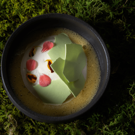 some kind of dessert: yellow bubbly substance, white cream substance, green broken shards, pink and yellow petals, inside black bowl, on top of faux grass