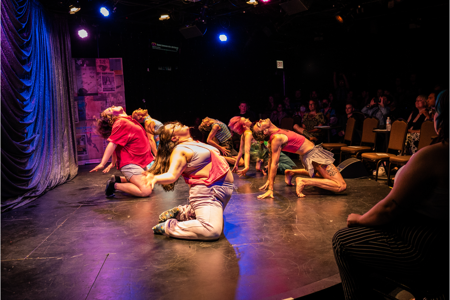 dancers in t-shirts or tank tops on their knees, some barefoot, others with socks on, audience watching, blue and purple glittery curtain