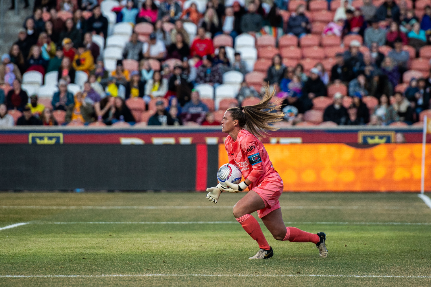 female soccer player in red uniform holding ball, blond hair in the air. audience watching